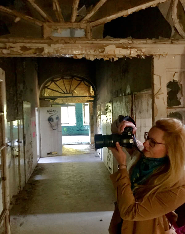 Eine Person mit Kamera steht in einem verlassenen, heruntergekommenen Gebäude mit abblätternden Wänden und einem offenen Torbogen im Hintergrund.