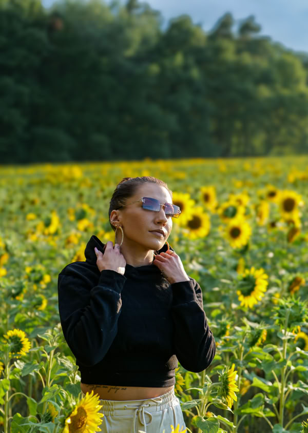 Eine Person mit Sonnenbrille steht in einem Sonnenblumenfeld, umgeben von blühenden Sonnenblumen, mit Bäumen im Hintergrund.