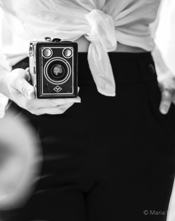 Eine Person hält eine alte Kamera in der Hand, trägt eine geknotete Bluse und eine dunkle Hose.