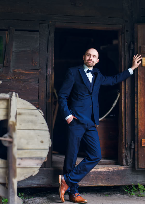 Ein Mann in einem blauen Anzug steht lässig an der Tür eines rustikalen Holzgebäudes.