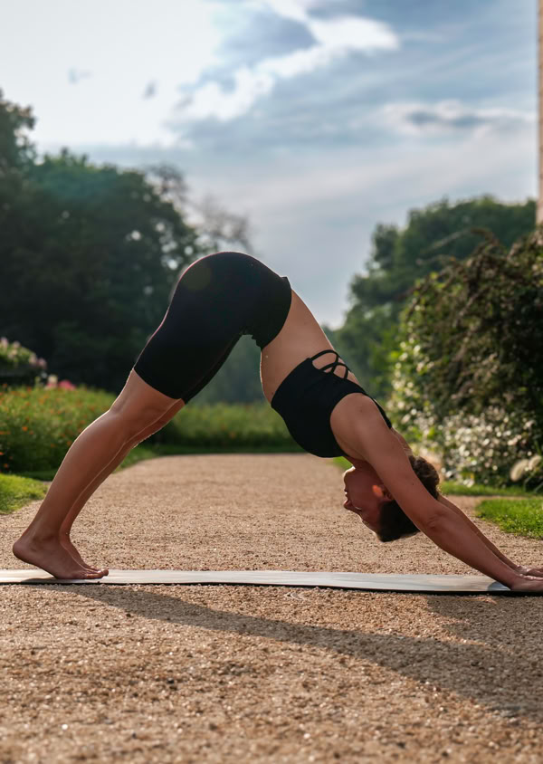 Person in schwarzer Sportkleidung macht eine Yoga-Pose im Freien auf einem Kiesweg, umgeben von Pflanzen und einem Gebäude im Hintergrund.