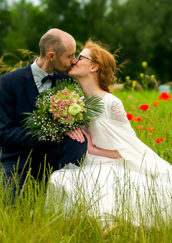 Ein Paar sitzt in einem blühenden Feld, die Frau trägt ein weißes Kleid und hält einen Blumenstrauß, während sie sich küssen.