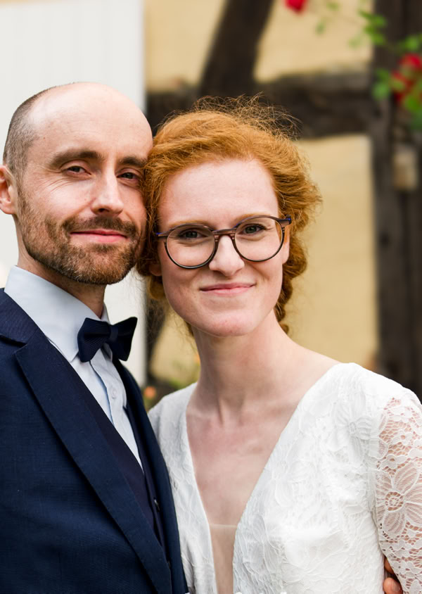 Ein Mann im Anzug und eine Frau im weißen Kleid stehen lächelnd vor einem Fachwerkhaus mit geöffnetem Fenster.