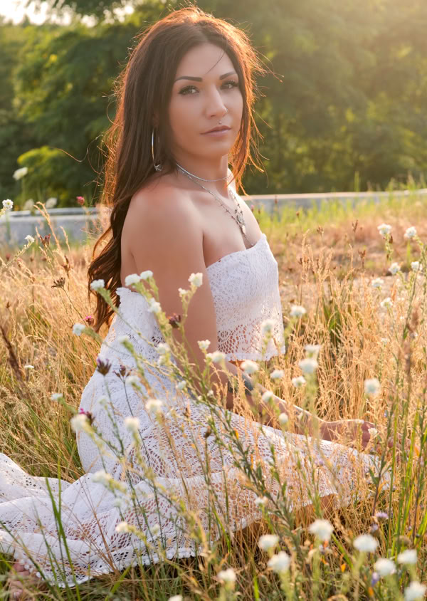 Frau in einem weißen Kleid sitzt im Gras auf einer Wiese bei Sonnenuntergang.