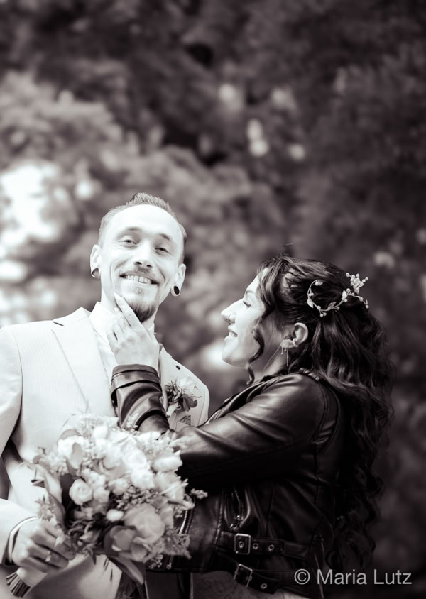 Ein lächelnder Mann im Anzug hält einen Blumenstrauß, während eine Frau in einer Lederjacke ihm liebevoll ins Gesicht fasst; im Hintergrund sind Bäume und ein Gebäude zu sehen.