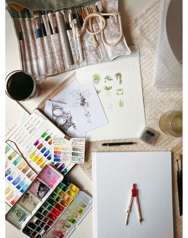 Künstlerbedarf auf einem Tisch, darunter Pinsel, Aquarellfarben, ein Skizzenbuch, ein Kompass und eine Tasse.