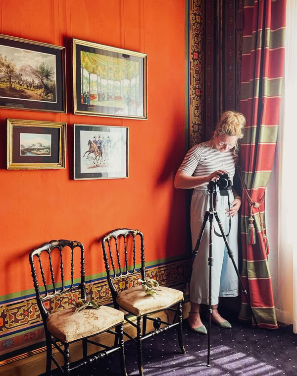 Eine Person steht neben einem Stativ in einem Raum mit orangefarbener Wand, an der mehrere Bilder hängen. Zwei Stühle stehen an der Wand, und ein großes Fenster mit Vorhängen lässt Licht herein.