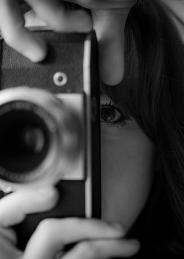 Schwarz-weiß-Foto einer Person, die eine Kamera vor ihrem Gesicht hält, mit einem Auge sichtbar durch den Sucher.